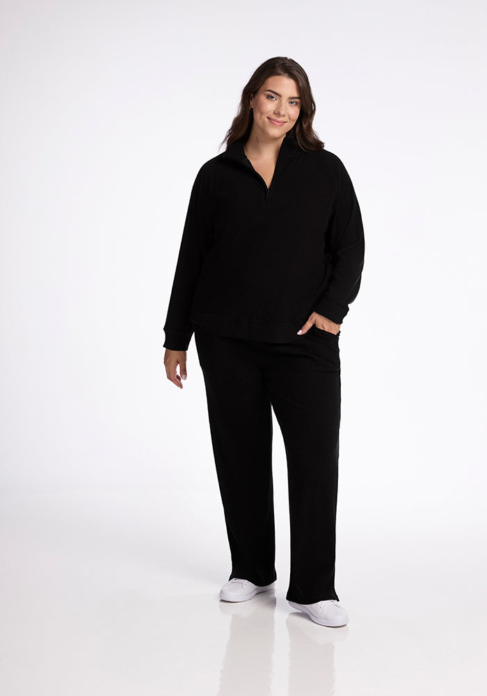 A person wearing the Woolx Quinn Ribbed 1/4 Zip in black paired with matching black pants stands against a plain white background. They have one hand in a pocket and are wearing white slip-on shoes, smiling slightly.