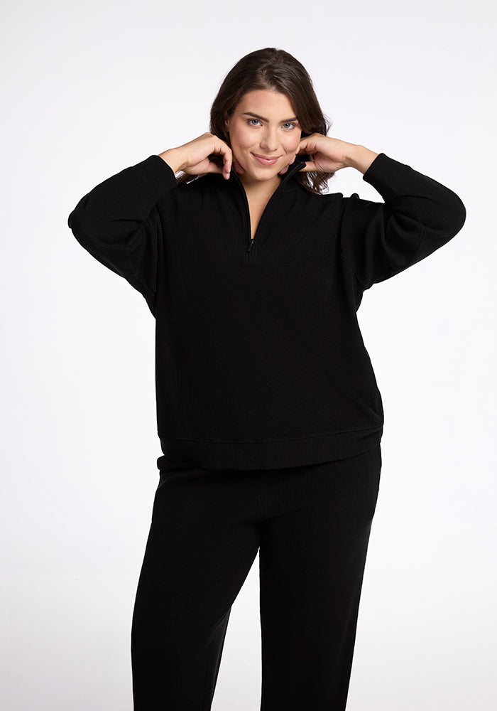 A person with long brown hair smiles as they adjust the collar of a Woolx Quinn Ribbed 1/4 Zip in black. They are wearing matching black pants and standing against a plain white background.