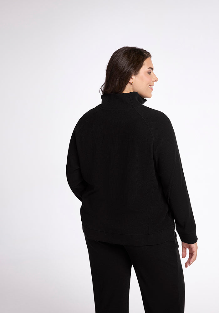 A person with long dark hair is wearing the Woolx Quinn Ribbed 1/4 Zip in black under a sleek jacket and pants, standing in a relaxed pose with a slight smile, facing away from the camera against a plain white background.