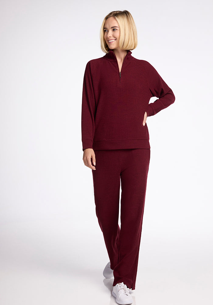 A person with short blonde hair is smiling and wearing the Quinn Ribbed 1/4 Zip in Cranberry Melange from Woolx paired with matching pants. They stand confidently, one hand in their pocket and the other on their hip, exuding comfort and style in white sneakers.