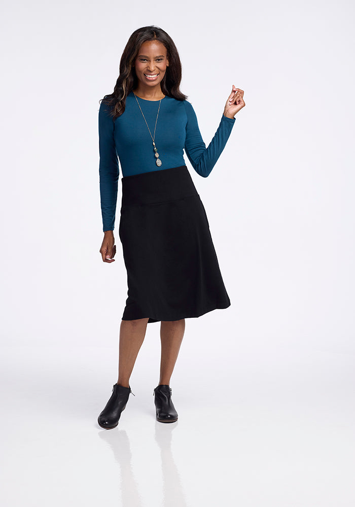 A woman smiles on a white background, wearing a teal long-sleeve top paired with the Marilyn Flowy Midi Skirt in black from Woolx. She accessorizes with a long necklace and black ankle boots. Her right arm is slightly raised to highlight the elegant, flowy design of her outfit.