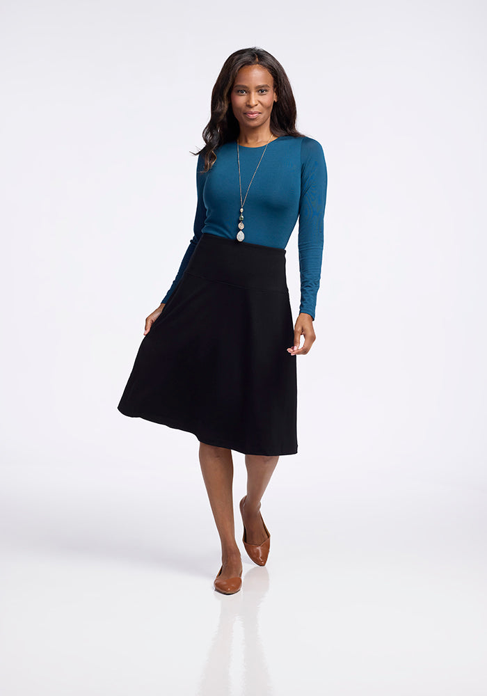 A woman strides confidently in a teal long-sleeve top and a Marilyn Flowy Midi Skirt in black from Woolx, featuring a chic flowy design. She elegantly complements her outfit with brown shoes and a long necklace adorned with multiple pendants, set against a clean white background that highlights her stylish ensemble.