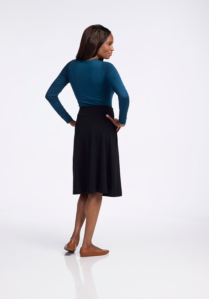 A person stands with their back to the camera, wearing a long-sleeved teal top, a Woolx Marilyn Flowy Midi Skirt in black made from merino wool, and brown shoes, set against a plain white background.