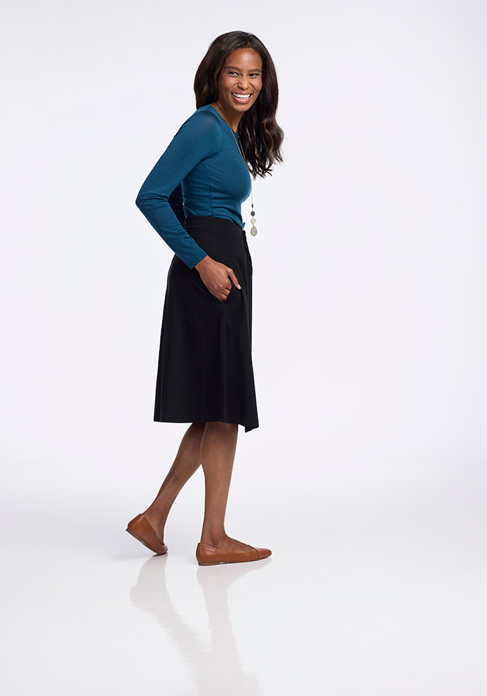 A woman stands sideways, smiling at the camera in a long-sleeve blue top and a Marilyn Flowy Midi Skirt - Black from Woolx, made of Merino wool, paired with brown flats. Her left hand rests casually in her skirt pocket against a plain white background.