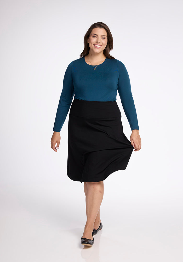 A person wearing a blue long-sleeve top and the Marilyn Flowy Midi Skirt in Black by Woolx walks confidently in a well-lit studio. They have long dark hair and are wearing black flats. The plain white background highlights the skirt's flowy design.