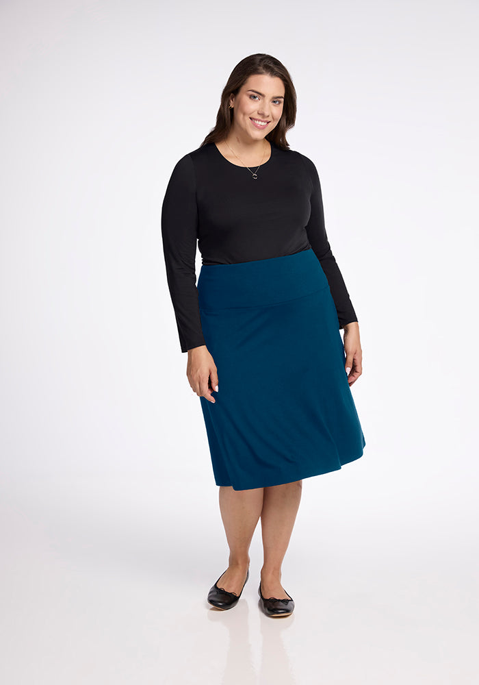 A woman stands smiling, dressed in a black long-sleeve top and the Marilyn Flowy Midi Skirt in Real Teal from Woolx, paired with black ballet flats against a plain white background.