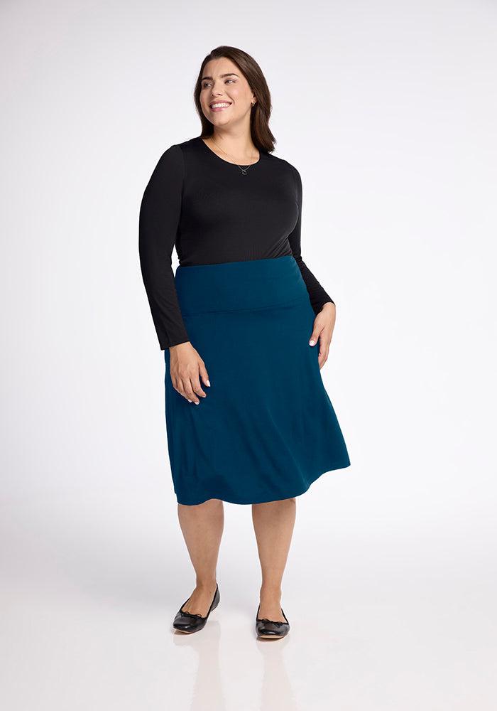 A person stands smiling against a plain background, wearing a black long-sleeve top paired with the Marilyn Flowy Midi Skirt in Real Teal by Woolx, styled in an elegant A-Line design and completed with black flat shoes.