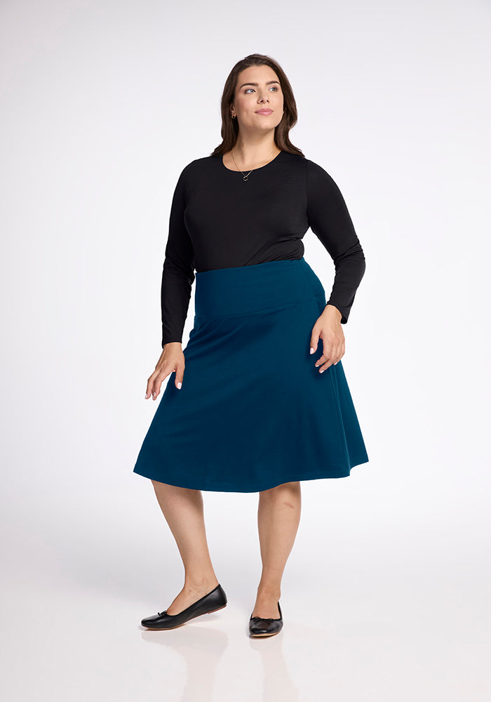 A woman wearing a black long-sleeve top, Marilyn Flowy Midi Skirt in Real Teal by Woolx with an A-Line design, and black flats poses against a plain white background. She has long hair and stands with one leg slightly forward, exuding effortless elegance.