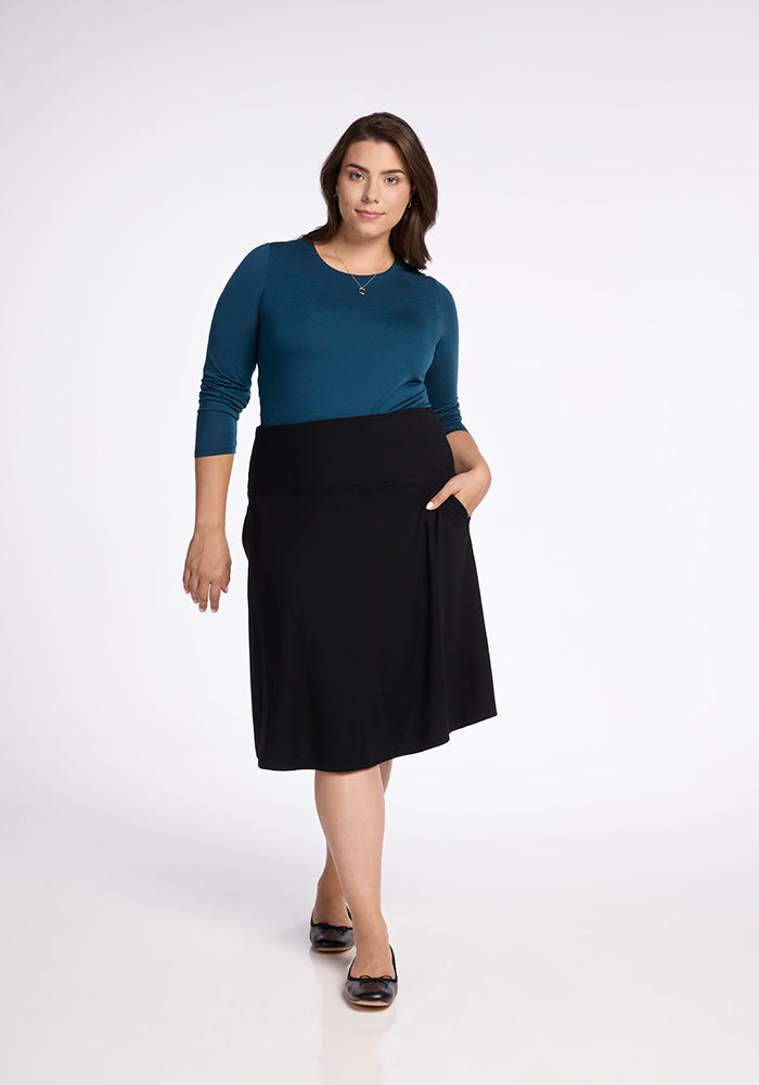 A woman exudes confidence in a teal long-sleeve top, beautifully paired with the Woolx Marilyn Flowy Midi Skirt in black. One hand rests casually in her pocket as she gazes forward with a neutral expression, her ensemble elegantly completed by black flats against the minimalist white backdrop.