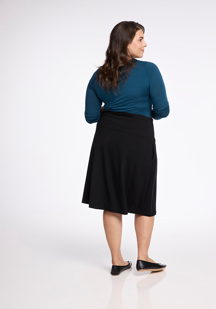 A woman with long brown hair stands with her back to the camera. She is wearing a teal top, a flowy Marilyn Flowy Midi Skirt by Woolx in black, and black flats. The background is plain white.