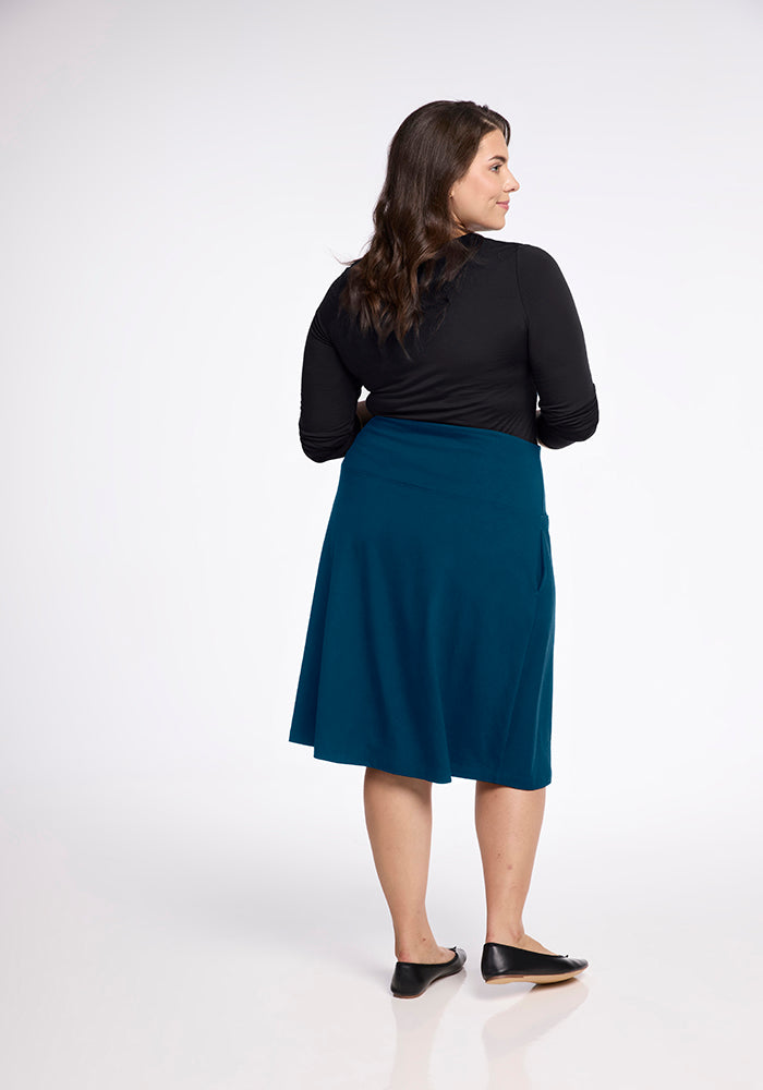 A woman with long brown hair, dressed in a black top and the Woolx Marilyn Flowy Midi Skirt in Real Teal with an A-line design, stands facing away and slightly to the side. Her arms are crossed, and she wears black flats. The background is plain white.