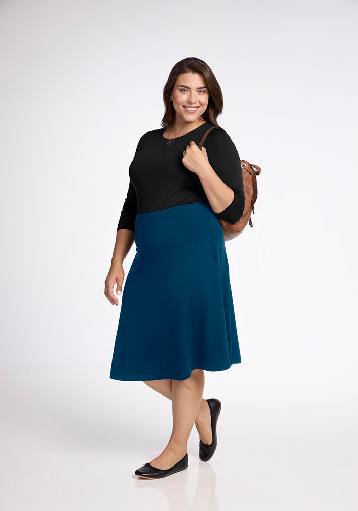 A woman with shoulder-length brown hair smiles warmly, wearing a black long-sleeve top paired with the Marilyn Flowy Midi Skirt in Real Teal from Woolx. She completes her outfit with black flats and carries a brown backpack over one shoulder. The plain white background emphasizes her chic style and the skirt's subtle A-line design.