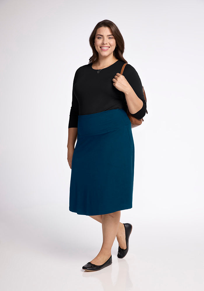 A person is smiling against a white background, dressed in a black long-sleeve top and the Marilyn Flowy Midi Skirt in Real Teal by Woolx, featuring an A-Line design. Completing the look are black flats and a brown backpack on their shoulder.