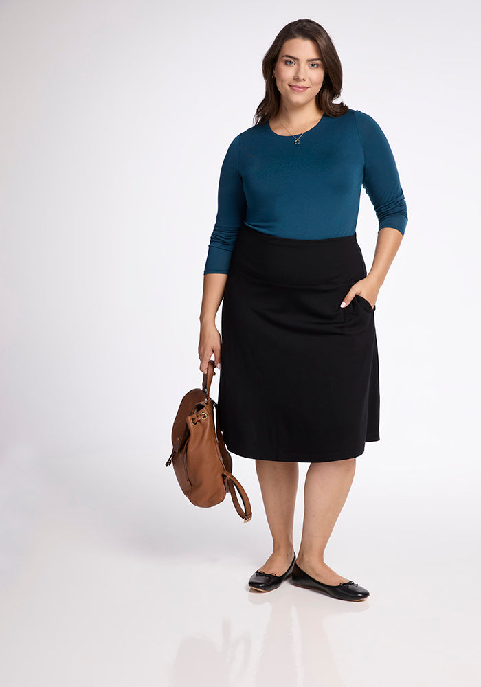 In front of a simple backdrop, a woman poses wearing a teal long-sleeve top and the Woolx Marilyn Flowy Midi Skirt in Black. She carries a brown backpack and completes her look with black flats made from merino wool, smiling gently at the camera.