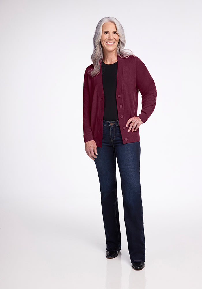 A woman with long gray hair stands smiling, wearing the Gia Sweater in Cabernet Crush by Woolx over a black top and dark jeans. The background is plain and white.