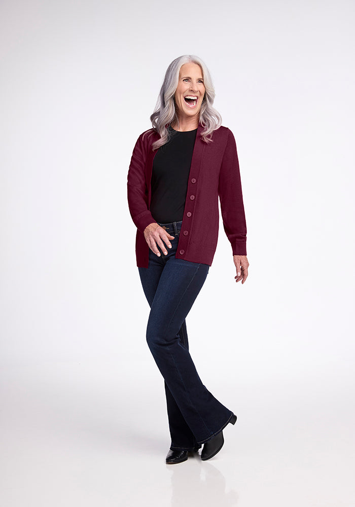 A woman with long gray hair, wearing the Woolx Gia Sweater in Cabernet Crush, a black top, and blue jeans, stands laughing against a plain background. She has one foot forward and appears joyful.