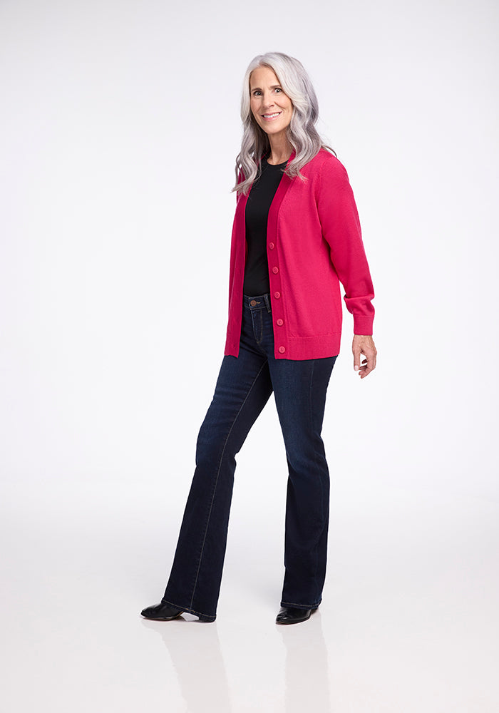 A woman with long gray hair, wearing a black top, the Gia Sweater in Berry Burst by Woolx, and dark blue jeans, stands smiling against a light background. She is facing slightly to the side with one hand in her pocket.