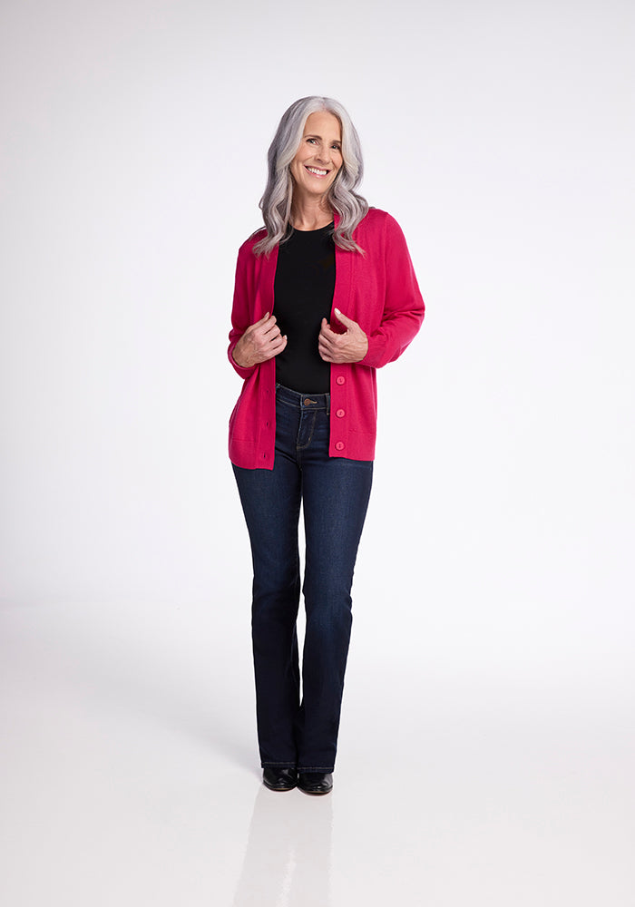 A person with long gray hair is smiling and standing against a plain white background. They are wearing the Woolx Gia Sweater in Berry Burst, a black top, dark blue jeans, and black shoes. They have their hands holding the edges of the unbuttoned Gia Sweater.