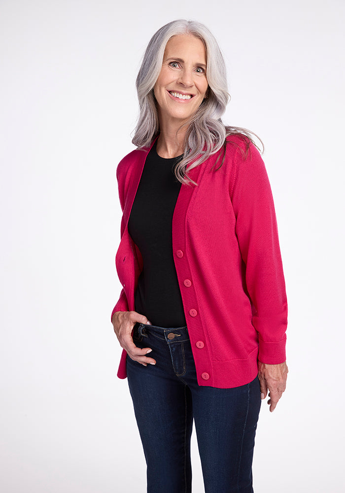 A smiling woman with long gray hair is wearing the Woolx Gia Sweater in Berry Burst over a black top and dark jeans. She stands against a plain white background with her hand in her pocket and the cardigan open. 