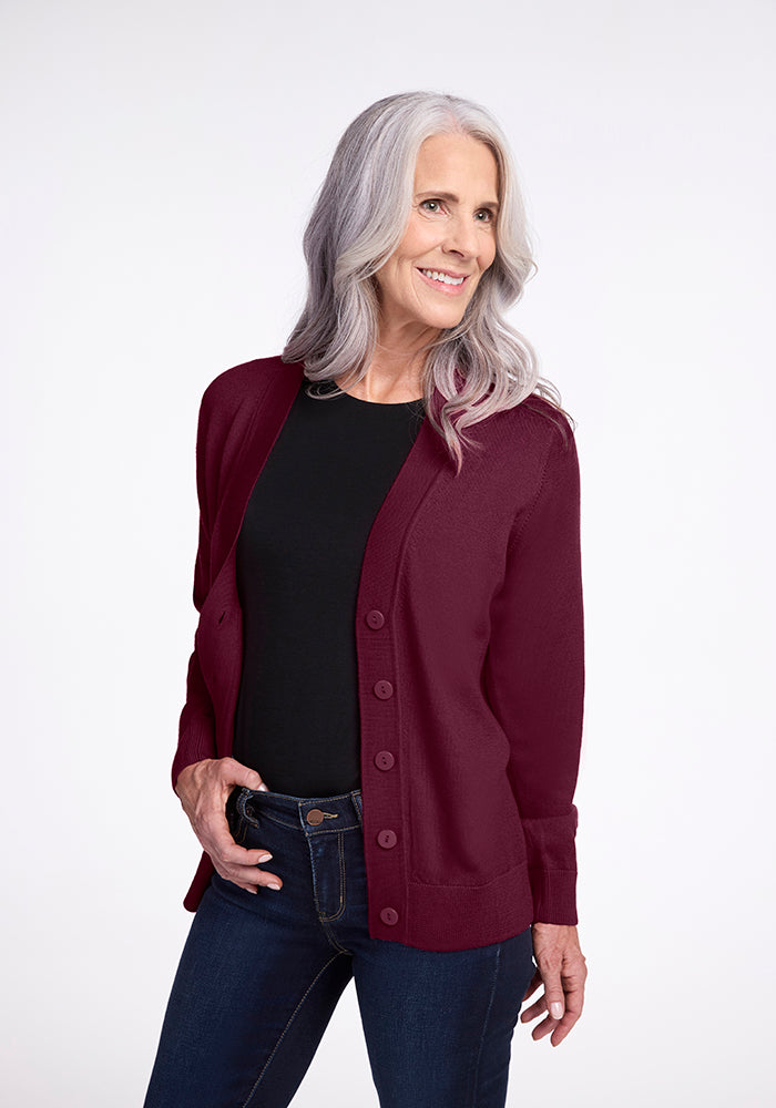 A smiling woman with long gray hair wears the Gia Sweater in Cabernet Crush from Woolx over a black shirt with jeans. She stands confidently against a plain white background.