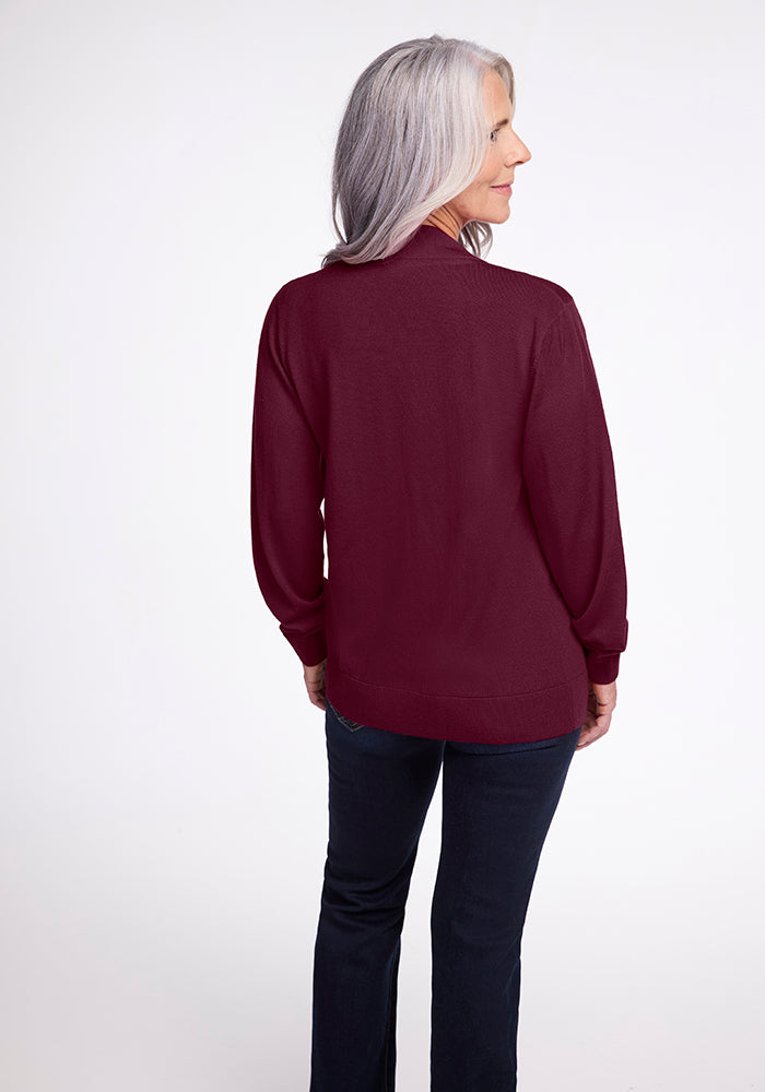 A person with long gray hair stands facing away, wearing the Gia Sweater in Cabernet Crush from Woolx along with dark jeans. They are on a plain white background, slightly turned to the side, smiling.