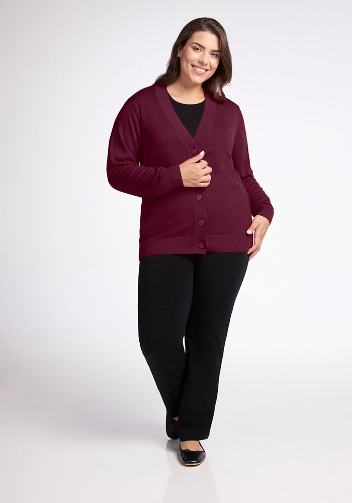 A person smiling and wearing the Woolx Gia Sweater - Cabernet Crush over a black top with black pants and shoes, standing against a plain white background.