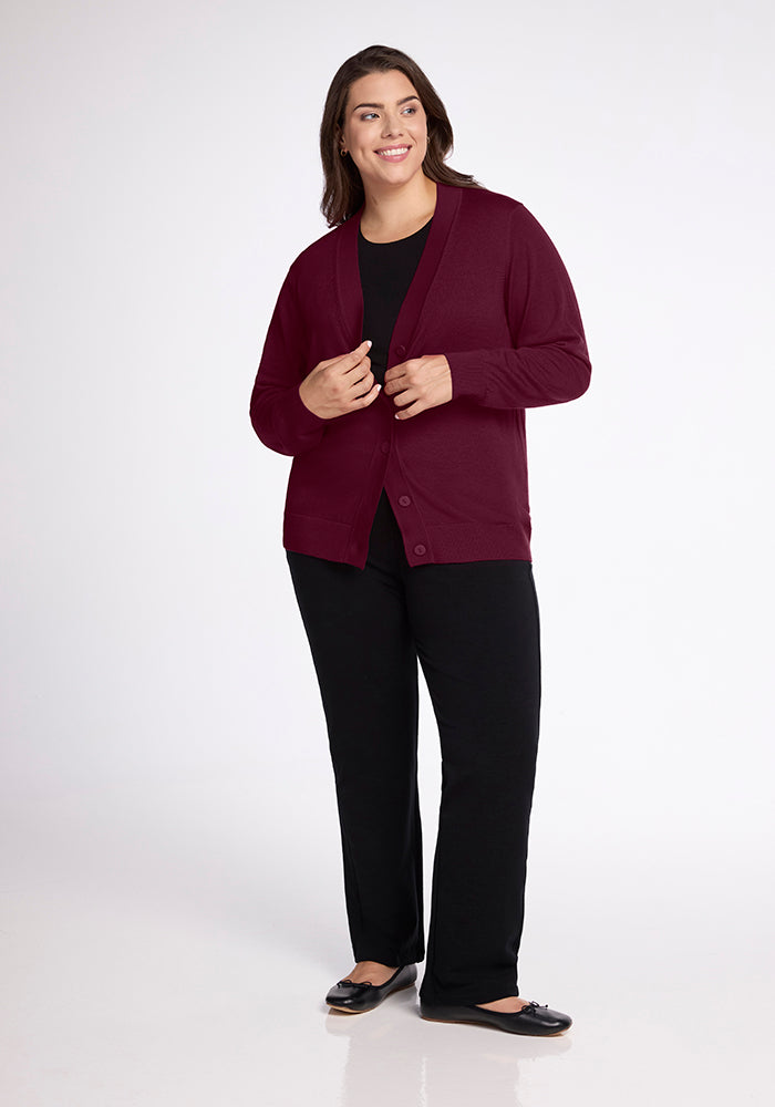 A person wearing the Gia Sweater in Cabernet Crush from Woolx over a black top and black pants smiles while looking off to the side. They are standing against a plain white background.