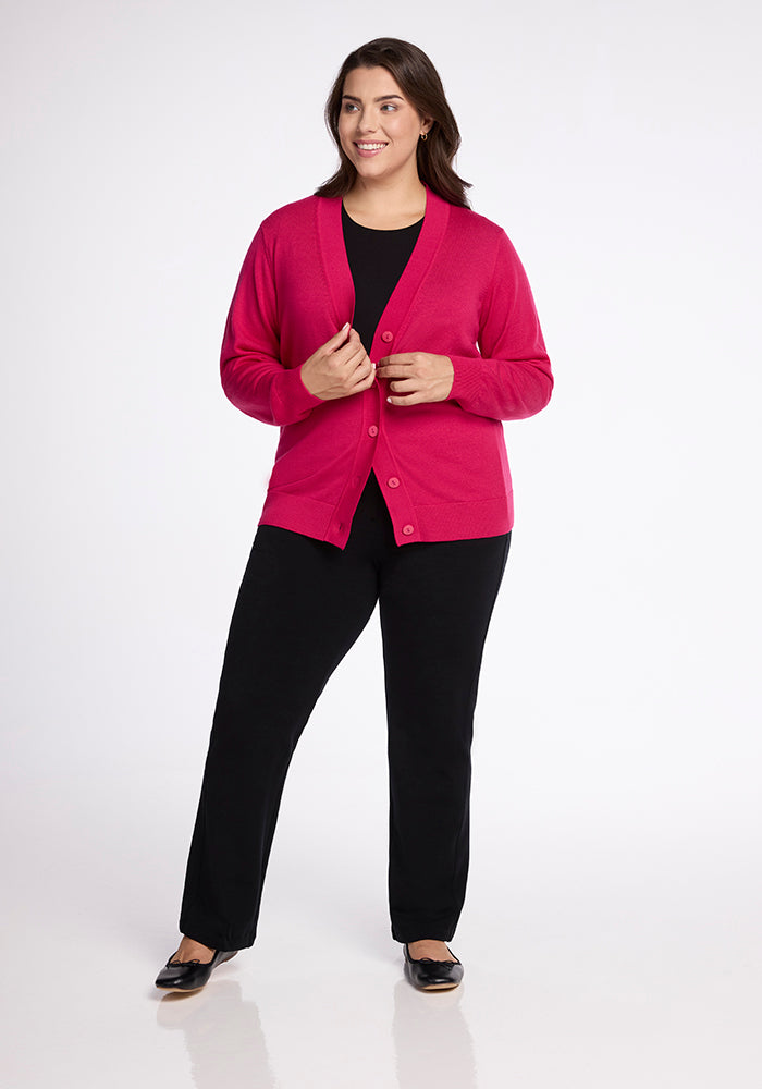 A woman stands smiling with her hands near the buttons of her Gia Sweater in Berry Burst, from the brand Woolx. She is also wearing a black top and black pants, along with black shoes. The background is solid white.