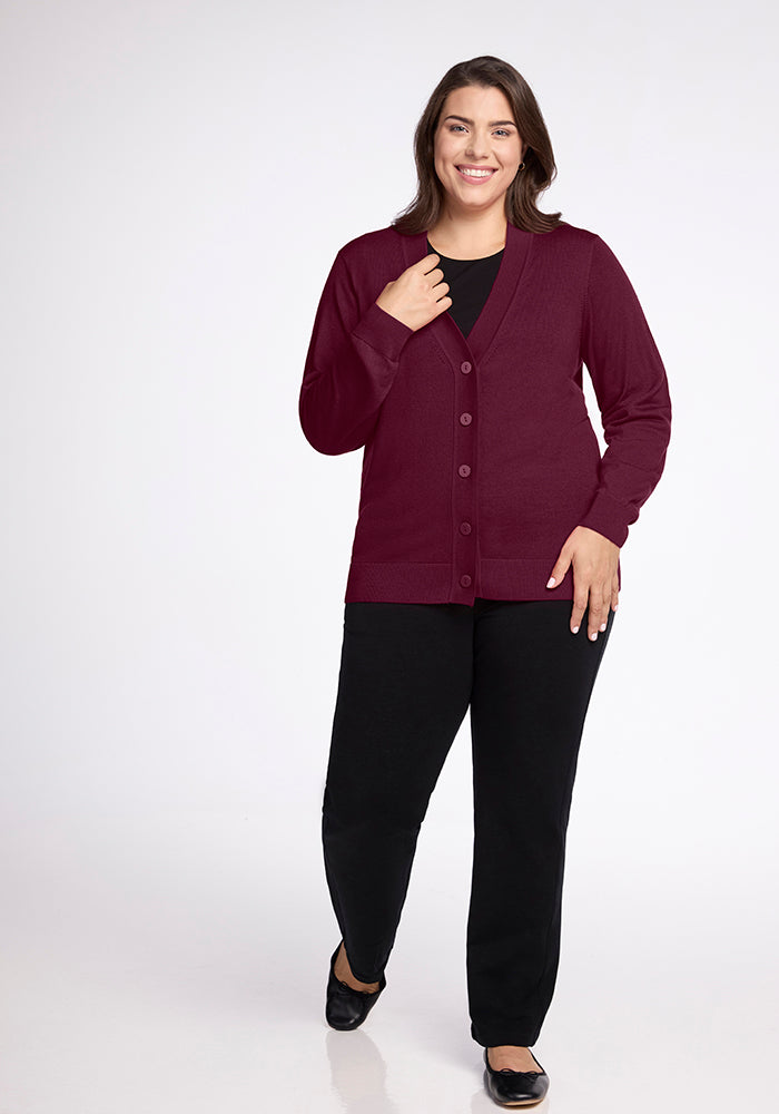 A person with long hair is smiling and walking towards the camera, wearing the Gia Sweater in Cabernet Crush from Woolx over a black top and trousers. They are standing against a plain white background.