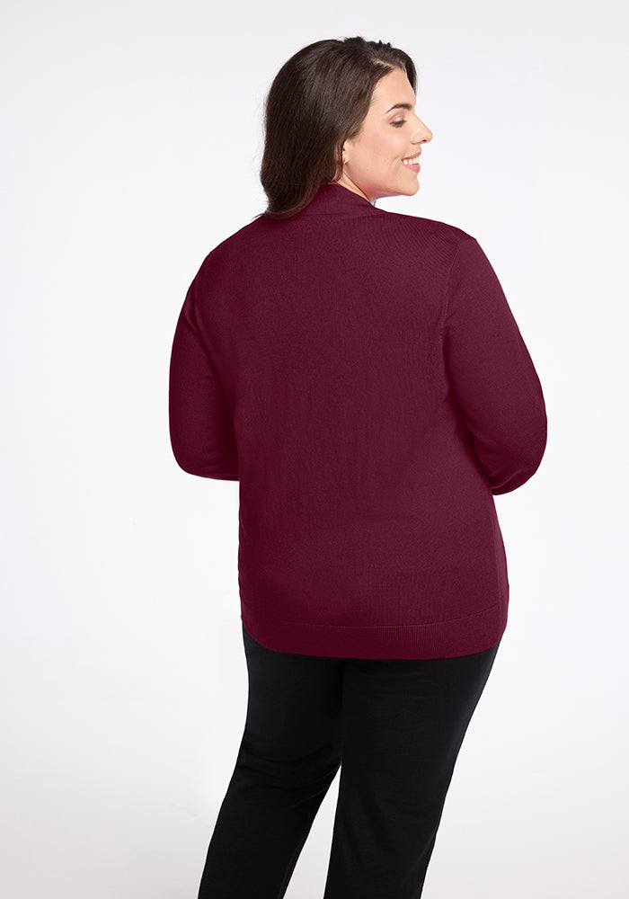 A woman with long dark hair is standing and smiling, wearing the Gia Sweater in Cabernet Crush by Woolx paired with black pants. She is facing away from the camera against a plain white background.