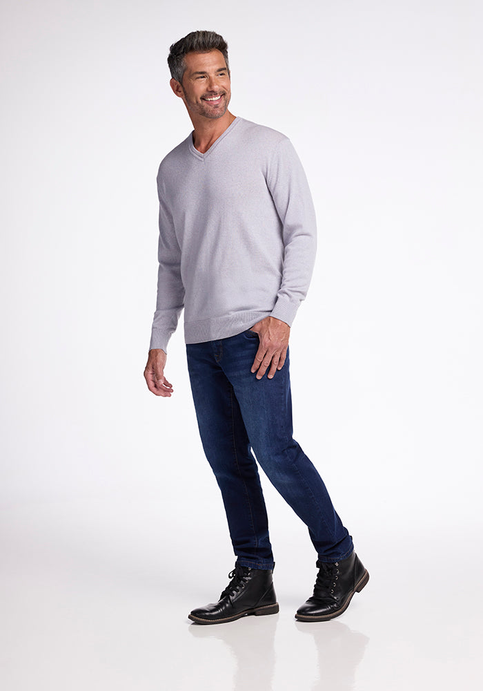 Man smiling and standing on a reflective surface, wearing a Mist-colored Vincenzo Sweater by Woolx, dark blue jeans, and black boots. The background is plain white.