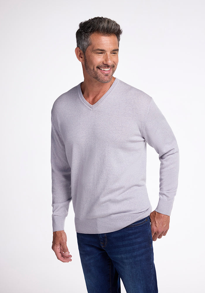 A smiling man wearing the Woolx Vincenzo Sweater in mist and dark blue jeans stands against a plain white background. His hair is short and graying, and he has a trimmed beard.