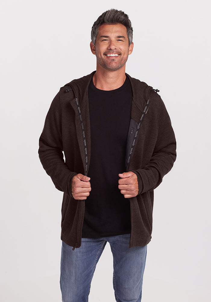 A man with short, greying hair smiles while standing against a plain white background. He is wearing a black t-shirt, jeans, and an unzipped dark brown Woolx Grizzly Sweatshirt in Maple Melange made of ultra-heavyweight Australian Merino Wool. He has his hands lightly holding the sweatshirt edges.