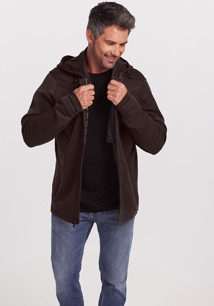 A man with short, graying hair is smiling while adjusting the collar of his dark Grizzly Sweatshirt - Maple Melange by Woolx. He is wearing a black t-shirt underneath and light blue jeans. The background is plain and light-colored.