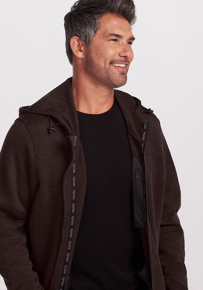 A smiling man with short, dark hair streaked with gray, is wearing a dark brown Woolx Grizzly Sweatshirt - Maple Melange over a black shirt. The sweatshirt has the zipper partially open, revealing a glimpse of an ultra-heavyweight black inner pocket on the left side. The background is plain white.