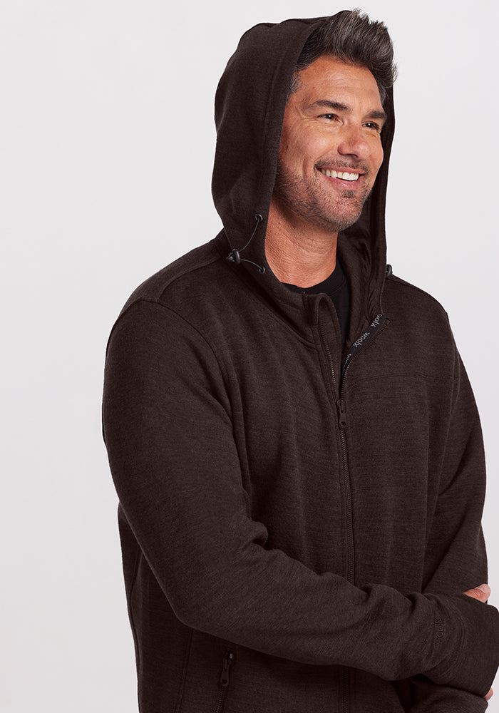 A man with short, dark hair smiles while wearing an ultra-heavyweight dark brown Woolx Grizzly Sweatshirt in Maple Melange made from Australian Merino Wool, with the hood up. The background is plain and light-colored. His arms are crossed in a relaxed pose.