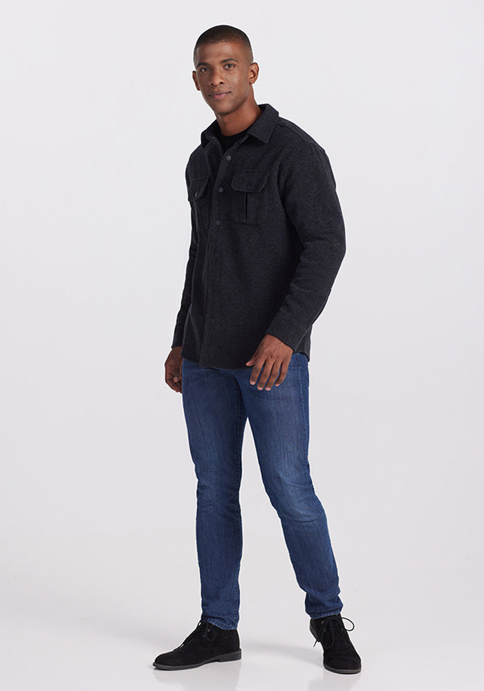 A man standing and facing slightly towards the left. He is wearing a ultra-functional Wilder Shirt Jac - Carbon Black from Woolx, which is made of Merino wool with buttoned pockets on the chest, blue jeans, and black shoes. The background is plain white.