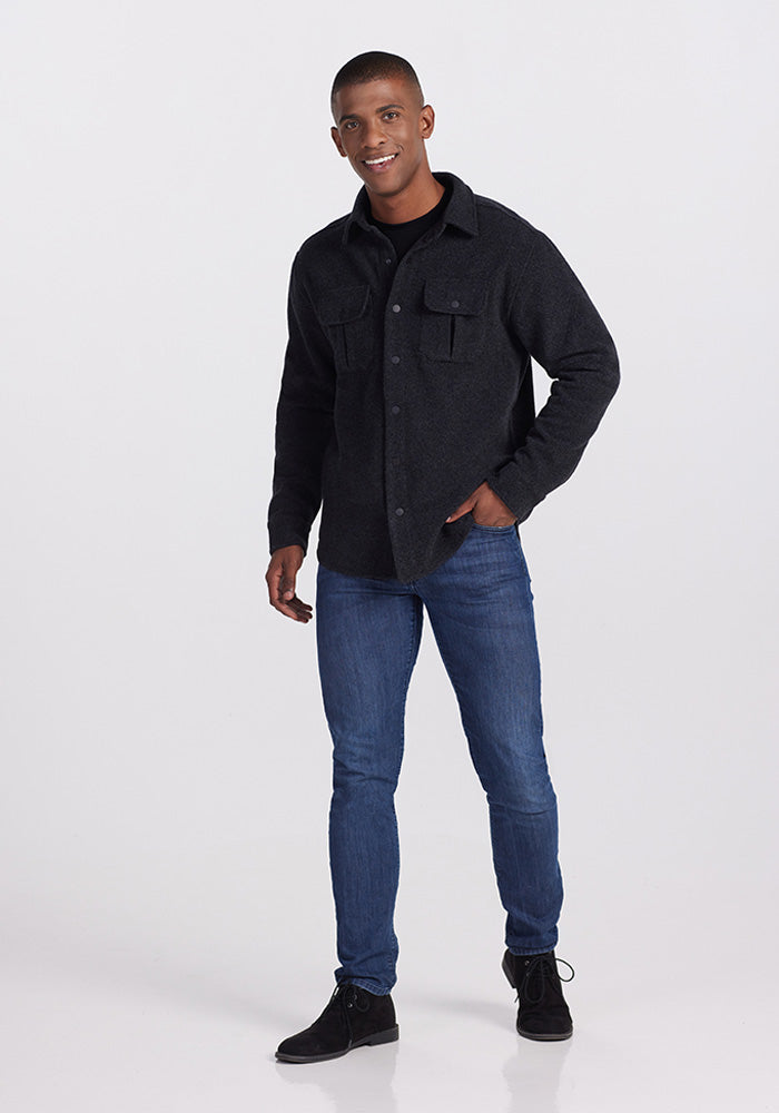 A person with short hair is standing and smiling, wearing the ultra-functional Woolx Wilder Shirt Jac in Carbon Black, blue jeans, and black shoes. They are posing casually with one hand in a pocket and the other hanging by their side against a plain, light-colored background.