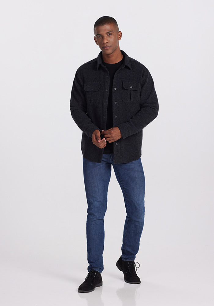 A man stands against a plain backdrop with his hands loosely clasped. He is wearing the Woolx Wilder Shirt Jac in Carbon Black over an ultra-functional Merino wool t-shirt, blue jeans, and black shoes. He is looking directly at the camera.