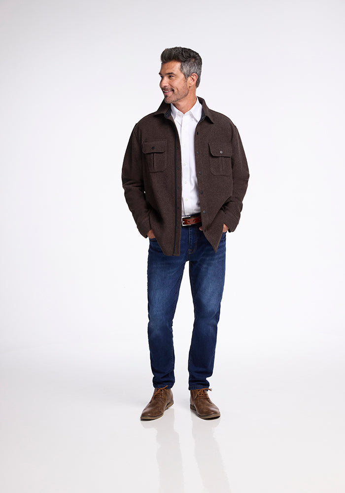 A man wearing the Woolx Wilder Shirt Jac over a white shirt, paired with blue jeans and brown shoes, stands against a white background. With his hands in his jacket pockets, he looks slightly to his left with a smile, showcasing an ultra-functional outfit that combines casual comfort with classic lumberjack style.