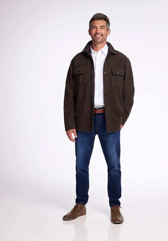 A man stands against a plain white background, smiling. He is wearing the ultra-functional Wilder Shirt Jac by Woolx in dark brown heavyweight fabric, paired with a white dress shirt, blue jeans, and brown shoes. His hands are in his jacket pockets.