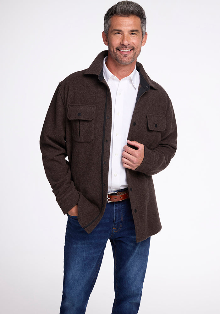 A man with short gray hair and a beard, exuding a lumberjack style, smiles while wearing the Woolx Wilder Shirt Jac in Maple Melange over a white shirt and blue jeans. He has his left hand in his pocket and his right hand holding the jacket near the top button. The background is plain white. 