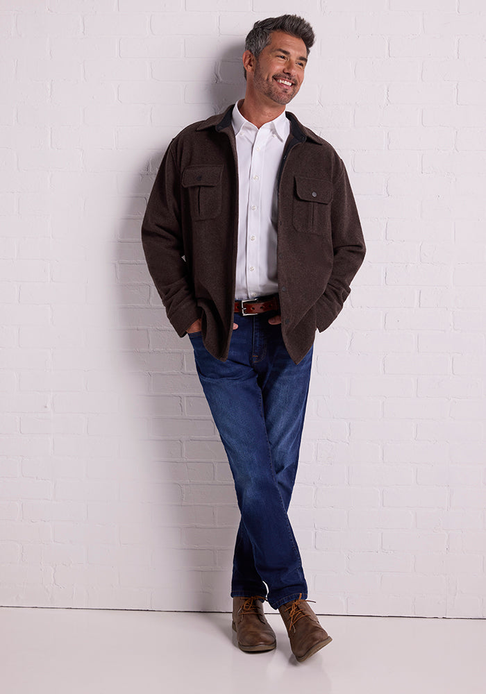 A man with gray hair and beard, sporting the Woolx Wilder Shirt Jac in Maple Melange over a white Merino wool shirt, blue jeans, and brown shoes, stands against a white brick wall. Embracing the lumberjack style, he is smiling with his hands in his jacket pockets and one leg crossed over the other.