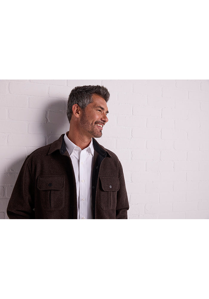 A man with short dark hair and a beard is looking to the side and smiling. He is wearing a white collared shirt under a dark brown, ultra-functional Wilder Shirt Jac by Woolx, featuring pockets, against a light-colored brick wall background.