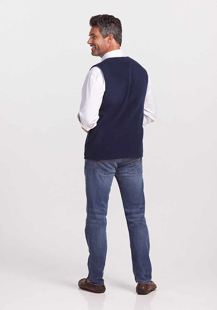 A man stands facing away, wearing a white shirt, the Woolx Baker Vest in Dark Navy, blue jeans, and brown shoes. He is smiling with his hands in his pockets.