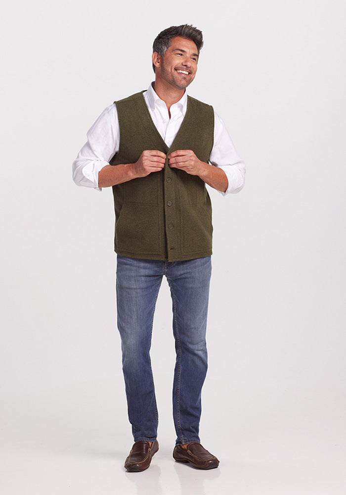 A smiling man stands against a plain background, wearing a white shirt, the Baker Vest in Dark Forest by Woolx, blue jeans, and brown shoes. He is adjusting his vest with his hands.