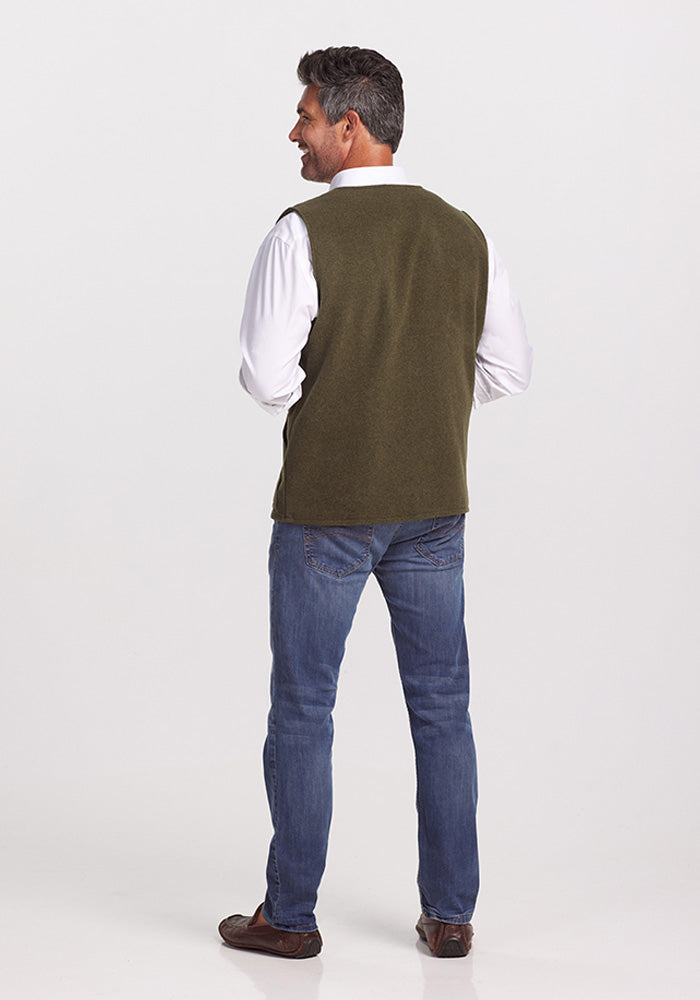 A man wearing the Woolx Baker Vest in Dark Forest and blue jeans stands facing away. He is smiling, has short dark hair, and is wearing a white shirt underneath the vest. He stands on a plain white background, embodying warmth and style.