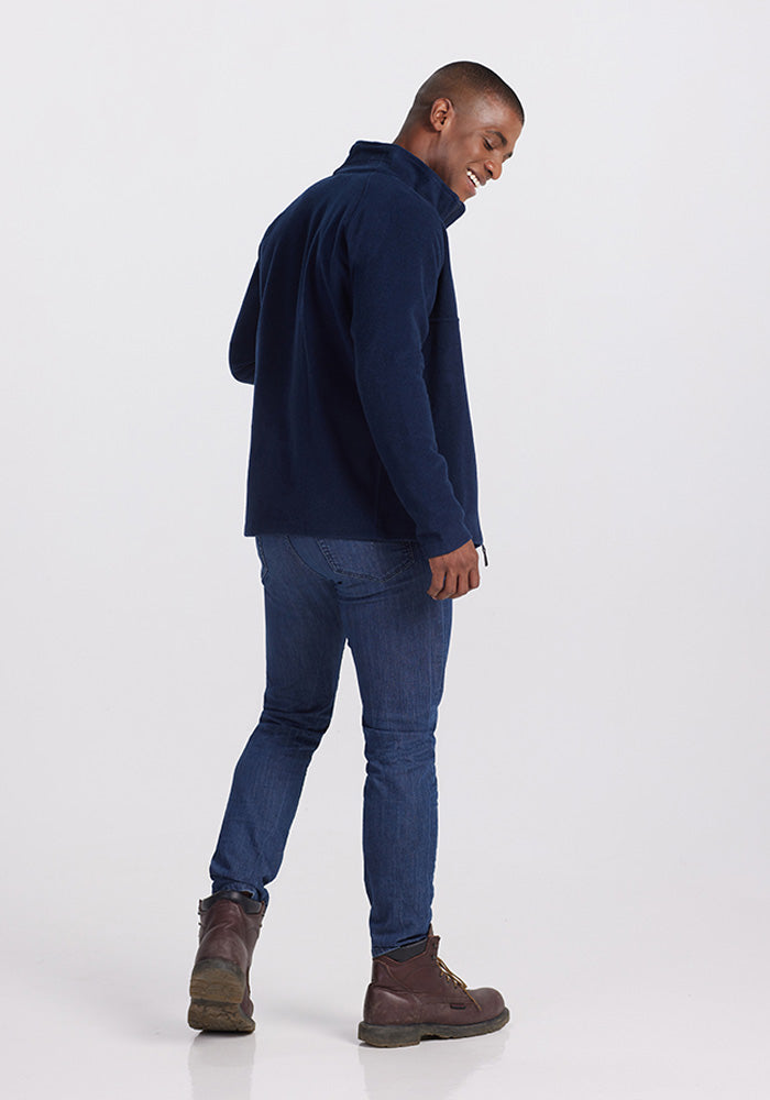 A person wearing a Woolx Fairbanks Jacket in Dark Navy, paired with blue jeans and brown boots, is standing and smiling. The individual is facing slightly sideways against a plain white background, giving a relaxed and casual pose.