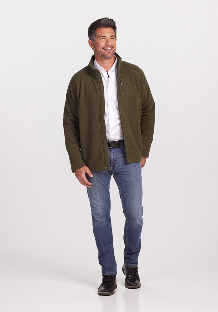 A man with short dark hair is smiling and walking, dressed in a Woolx Fairbanks Jacket - Dark Forest, celebrated for its insulation. He pairs it with a light gray Merino wool shirt, blue jeans, and black shoes against a plain white backdrop.