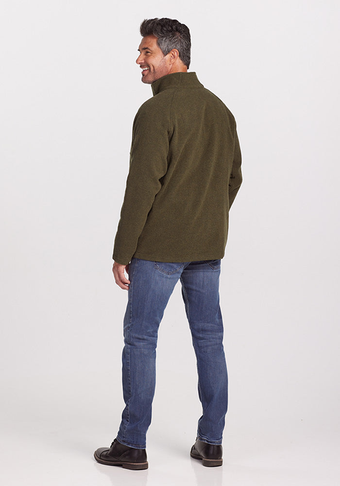 A man is standing and smiling, turned slightly to his left, wearing a stylish Fairbanks Jacket - Dark Forest in olive green by Woolx. He pairs it with blue jeans and black shoes, all against a plain white background that accentuates his relaxed demeanor.